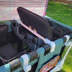 May include: A teal wagon with a black padded seat, cup holders, and storage pockets. The wagon has a pink and white patterned canopy and a white hook on the side. The wagon has a patch with the text "Every park deserves a good time".