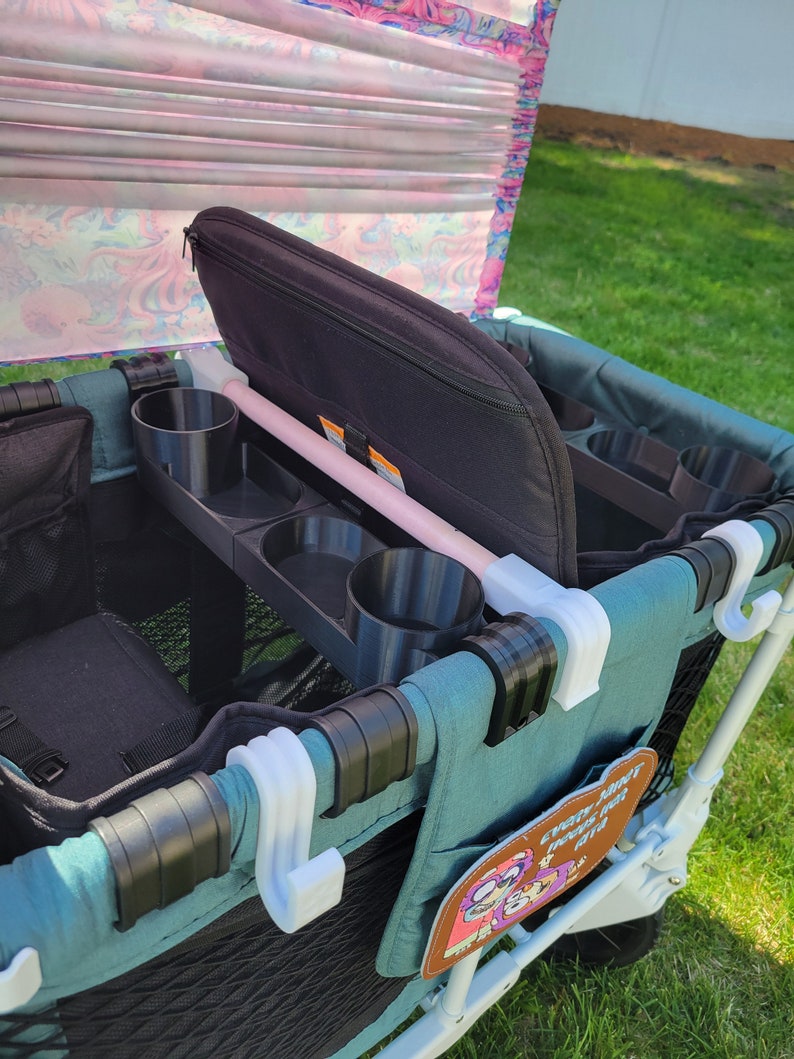 May include: A teal wagon with a black padded seat, cup holders, and storage pockets. The wagon has a pink and white patterned canopy and a white hook on the side. The wagon has a patch with the text "Every park deserves a good time".