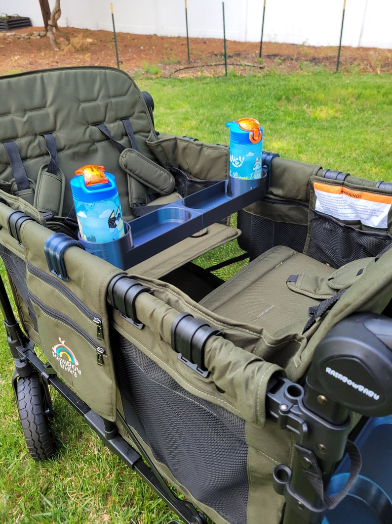 May include: A green wagon with two seats, a black handle, and a black tray with two cup holders. The wagon has a black mesh side and a black and white logo that says "Rainbow Buggy". There are two blue water bottles with orange lids in the cup holders.