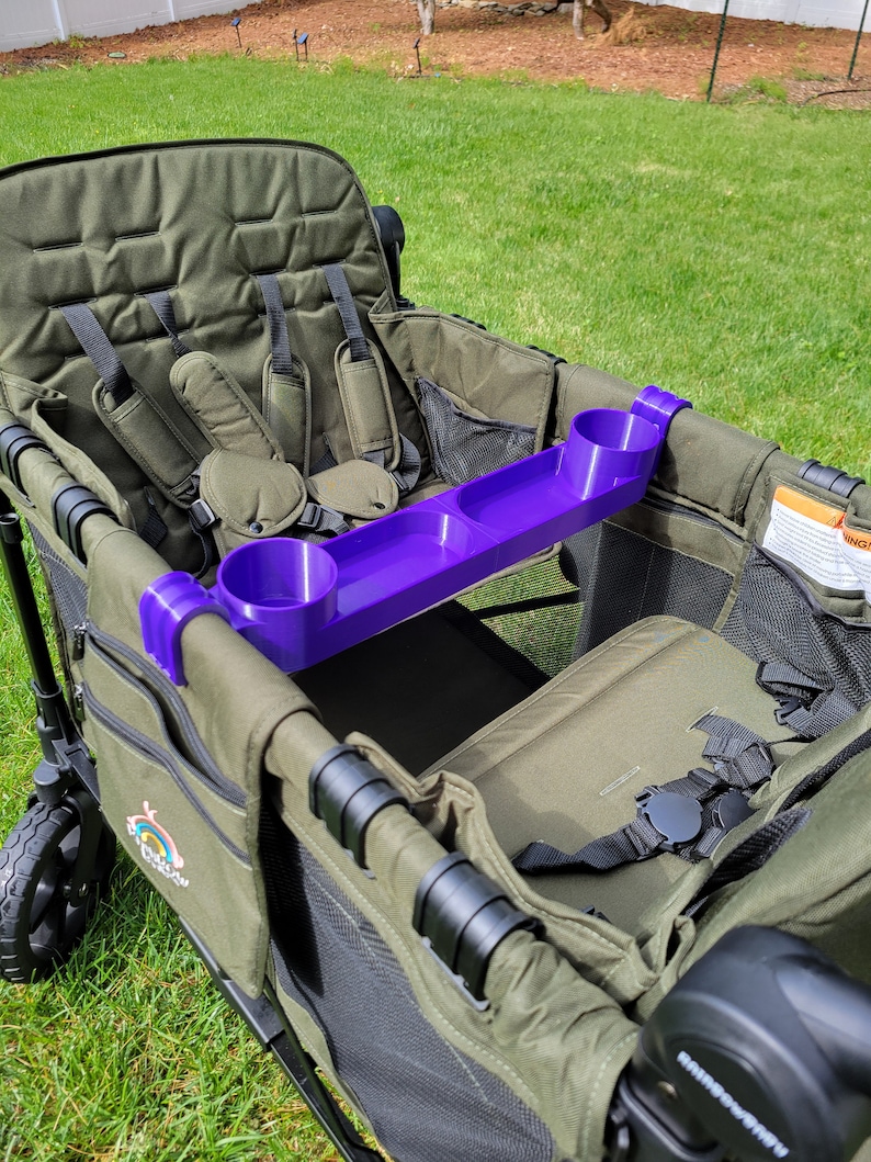 May include: A purple cup holder accessory for a wagon or stroller. The cup holder has three slots for cups or bottles and is attached to the side of the wagon.