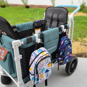 May include: A white wagon with a teal fabric interior and two black seats. Two dinosaur-themed backpacks are hanging from the wagon's hooks. One backpack is white with pink, blue, and yellow dinosaurs, and the other is blue with black, white, and yellow dinosaurs. The wagon has a black and white patch that says "CUTE, BUT BIG".