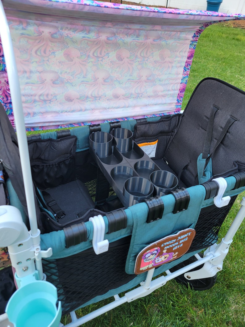 May include: A teal and white wagon with a black and gray cup holder. The wagon has a black mesh storage area and a black fabric seat. The wagon has a pink and white canopy with an octopus pattern. The wagon has a white hook on the side and a brown patch with the text "every adventure begins with a ride".