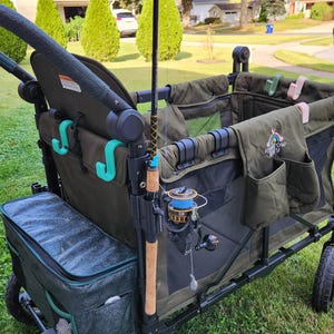 May include: A dark green utility wagon with a fishing rod attached. The wagon has a cooler, storage pockets, and a black frame. The fishing rod is brown and black with a blue and black reel. The wagon is on a grassy lawn.