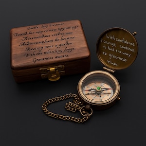 Peut inclure: Une boussole de style vintage avec un boîtier doré et une chaîne, à côté d'une boîte en bois. La boîte contient du texte gravé. La boussole est ouverte, révélant une citation à l'intérieur. Le fond est noir.