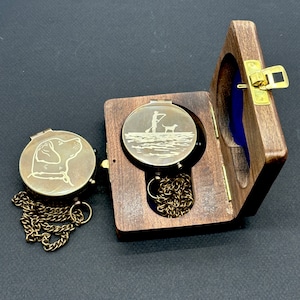 May include: Two brass compass pendants with chains. One pendant features a dog and the other features a person with a dog on a paddleboard. Both pendants are in wooden boxes.