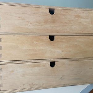 Könnte beinhalten: Eine Aufbewahrungseinheit aus hellem Holz mit drei vertikal gestapelten horizontalen Schubladen. Jede Schubladenfront besteht aus glattem Sperrholz mit sichtbarer Maserung und einem halbkreisförmigen Ausschnitt in der Mitte zum Öffnen. Der Rahmen zeigt Fingerzinken.