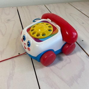 Puede incluir: Un teléfono de juguete para tirar Fisher-Price vintage. El juguete es blanco con un dial de arcoíris, un auricular rojo y ruedas rojas. Tiene detalles en azul y una cara sonriente. Una cuerda roja está adjunta.