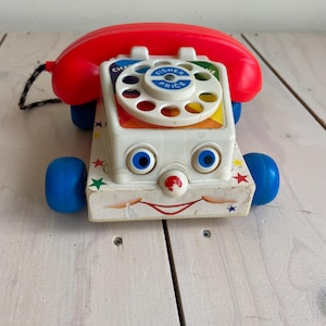 Puede incluir: Teléfono de juguete Fisher-Price Chatter Telephone vintage. El teléfono de juguete tiene un auricular rojo, un dial blanco y ruedas azules. El teléfono tiene una cara sonriente y decoraciones de estrellas. Un juguete clásico.