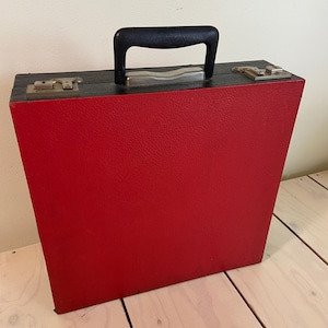Könnte beinhalten: Ein rechteckiger Tragekoffer mit einer strukturierten roten Außenseite und einer schwarzen Oberkante. Der Koffer verfügt über einen schwarzen Kunststoffgriff, zwei silberfarbene Metallverschlüsse an der Oberseite und ein Scharnier.