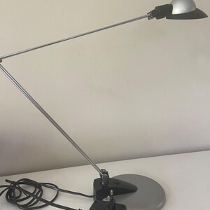 May include: A silver and black desk lamp with a flexible arm and a round base. The lamp has a black power cord and a black power adapter.