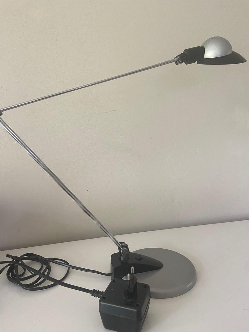 May include: A silver and black desk lamp with a flexible arm and a round base. The lamp has a black power cord and a black power adapter.