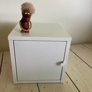 Peut inclure: Un meuble métallique blanc avec une seule porte et une petite poignée ronde. Une figurine en bois avec une tête floue et un collier en corde est posée sur le dessus du meuble. La figurine est marron avec des yeux blancs.