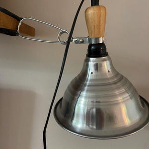 Könnte beinhalten: Eine Wandleuchte mit einem konischen Lampenschirm aus silberfarbenem Metall, einem hellen Holzgriff und einem schwarzen Stromkabel. Die Leuchte ist mit einer silberfarbenen Metall-Federklemme an einem hölzernen Tragarm befestigt.