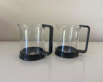 Op de afbeelding: Twee glazen bekers met zwarte kunststof handvatten en bodems. De bekers zijn leeg en staan op een wit oppervlak.