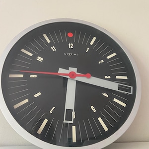 Op de afbeelding: Een ronde wandklok met een zilverkleurige rand en een zwarte wijzerplaat. De klok heeft witte cijfers van 1 tot 12, witte uurmarkeringen en de merknaam NEXTIME in het wit. De klok heeft een rode secondewijzer en twee brede, lichtgrijze wijzers.