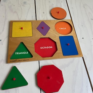 Può includere: Puzzle di forme in legno con una varietà di forme geometriche colorate, tra cui un quadrato giallo, un rombo viola e un triangolo verde. La parola "OCTAGON" e "CIRCLE" sono stampate sulla tavola. I pezzi del puzzle hanno pomelli rossi.