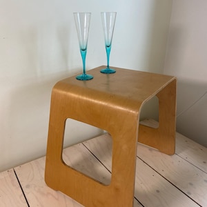 Puede incluir: Dos copas de champán altas y delgadas con cuencos de vidrio transparente y tallos y bases de color turquesa vibrante, colocadas sobre un taburete de madera clara curvada con un diseño de corte cuadrado en los paneles laterales y veta natural.