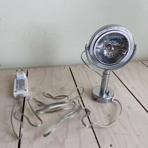 May include: A silver spotlight with a circular head and a stand. The spotlight has a bulb inside and is connected to a power adapter and cord. The spotlight is on a light-coloured wooden surface.