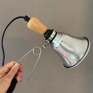 Puede incluir: Una lámpara de pinza de metal plateado con una pantalla en forma de campana con pequeños orificios de ventilación, un mango de madera clara y un cable eléctrico negro. El accesorio incluye un gran mecanismo de sujeción de metal con resorte.
