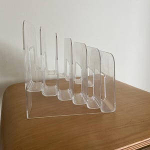 Pode incluir: Um organizador de acrílico transparente com seis divisórias verticais de alturas variáveis dispostas em formação escalonada. A estrutura transparente tem cantos superiores arredondados e uma base sólida para manter itens na vertical.