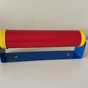 Könnte beinhalten: Eine Schreibtischlampe im Retro-Stil in Primärfarben. Die zylindrische Leuchte ist rot mit gelben Endkappen und wird von einem blauen, rechteckigen Sockel getragen. Die Lampe hat ein weißes Netzkabel.