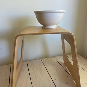 Puede incluir: Un taburete de madera clara con una parte superior curvada y asas recortadas. Un cuenco de cerámica blanca se encuentra encima. El taburete está hecho de madera contrachapada y tiene un diseño simple y moderno. El cuenco es redondo y tiene un borde ancho.