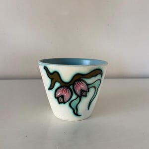 May include: A small, off-white ceramic cup with a light blue interior. The exterior features a floral design in pink, teal, and brown. The cup has a slightly flared shape and is likely vintage.