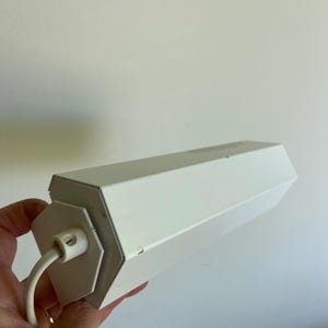 Può includere: Un apparecchio di illuminazione bianco di forma geometrica con un cavo bianco. L'apparecchio ha una forma esagonale e sembra essere in metallo. Il cavo è attaccato a un'estremità dell'apparecchio.