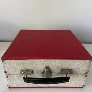 Pode incluir: Uma mala retangular vintage com uma parte superior vermelha texturizada e um corpo texturizado prateado e branco. A mala tem uma alça preta, fechos de metal e protetores de canto de metal. A mala está fechada.