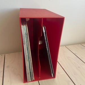 May include: A red vinyl record storage box with two compartments. The box is filled with vinyl records. The box is made of a textured material and is sitting on a light-colored wooden surface.