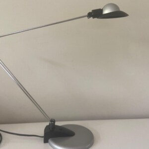 May include: A silver and black desk lamp with a flexible arm and a round shade. The lamp is on a white surface.
