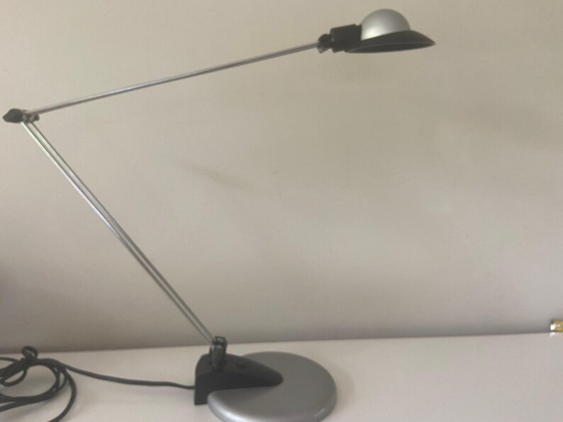 May include: A silver and black desk lamp with a flexible arm and a round shade. The lamp is on a white surface.