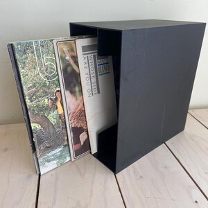 May include: A black vinyl record storage box with a slanted design. Several album covers are partially visible, including one with the text "WHOSE SIDE ARE YOU ON" and another with the artist name "MATT BLANCO". The box is on a light wood surface.