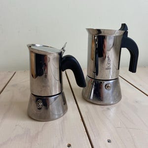 Könnte beinhalten: Eine weiße Keramik-Tasse mit schwarzem Henkel und dem Text "But First, Coffee" in fetter, schwarzer Schrift. Die Tasse steht vor einem neutralen Hintergrund, der ihr schlichtes Design hervorhebt.