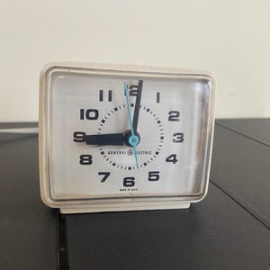 Puede incluir: Reloj despertador blanco con agujas y números negros. La esfera del reloj dice "General Electric" y "Made in U.S.A."