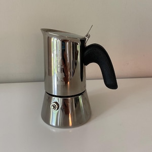 May include: A stainless steel stovetop espresso maker with a polished reflective finish. The cylindrical upper chamber features a small embossed logo of a person with a moustache. It has a black textured handle and a small metal pressure valve.