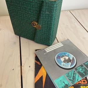 Könnte beinhalten: Ein grüner, texturierter Schallplattenkoffer mit einem messingfarbenen Metallverschluss. Daneben liegt eine quadratische Papierhülle mit einem blauen RAK-Plattenlabel in der Mitte sowie eine weitere Schallplattenhülle mit orangefarbenem und schwarzem Text.