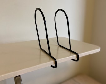 May include: Black metal bookends with a curved top design. The bookends are placed on a light beige surface. The minimalist design is suitable for organising books, magazines, or records.