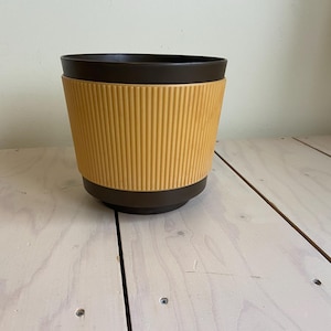 Puede incluir: Una maceta de dos tonos con la parte superior e inferior marrón y un centro acanalado de color amarillo mostaza. La maceta está hecha de un material duradero y es adecuada para uso en interiores o exteriores. El diseño es moderno y minimalista.