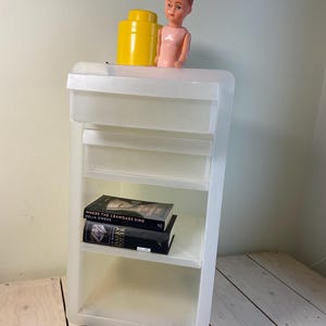 May include: A translucent white plastic rolling cart with three shelves and a drawer. A yellow container and a vintage doll sit on top. Two books are on the middle shelf, one titled "Where the Crawdads Sing".