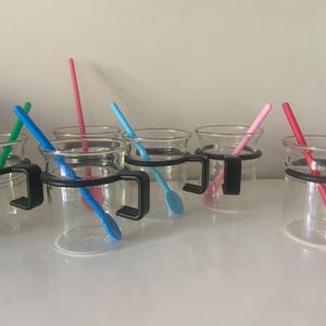 Op de afbeelding: Een set van vijf heldere glazen mokken, elk voorzien van een zwarte kunststof handgreep. In elke mok zit een lepel met een lange steel in een opvallende kleur: groen, blauw, roze, lichtblauw en rood. De mokken staan op een rij.