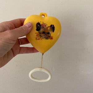 May include: A yellow heart-shaped plastic musical pull toy featuring a graphic illustration of a brown puppy with a red bow on its head. The toy includes a white plastic ring attached to a string at the bottom for pulling to activate the mechanism.