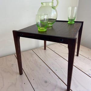 May include: A dark wood side table with a rectangular top and four legs. On the table are three green-tinted glass items: a jug and two glasses. The table is set on a light wood floor.