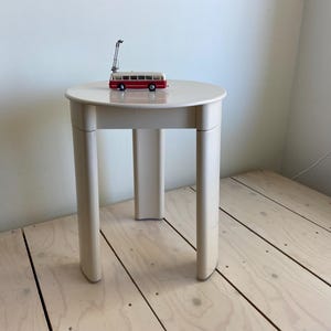 May include: A small, cream-colored side table with a round top and three legs. A miniature red and white bus toy with a black antenna sits on the table. The table is approximately 18 inches tall.