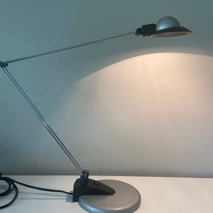 May include: A silver and black desk lamp with a flexible arm and a round base. The lamp is on and casting a warm light.