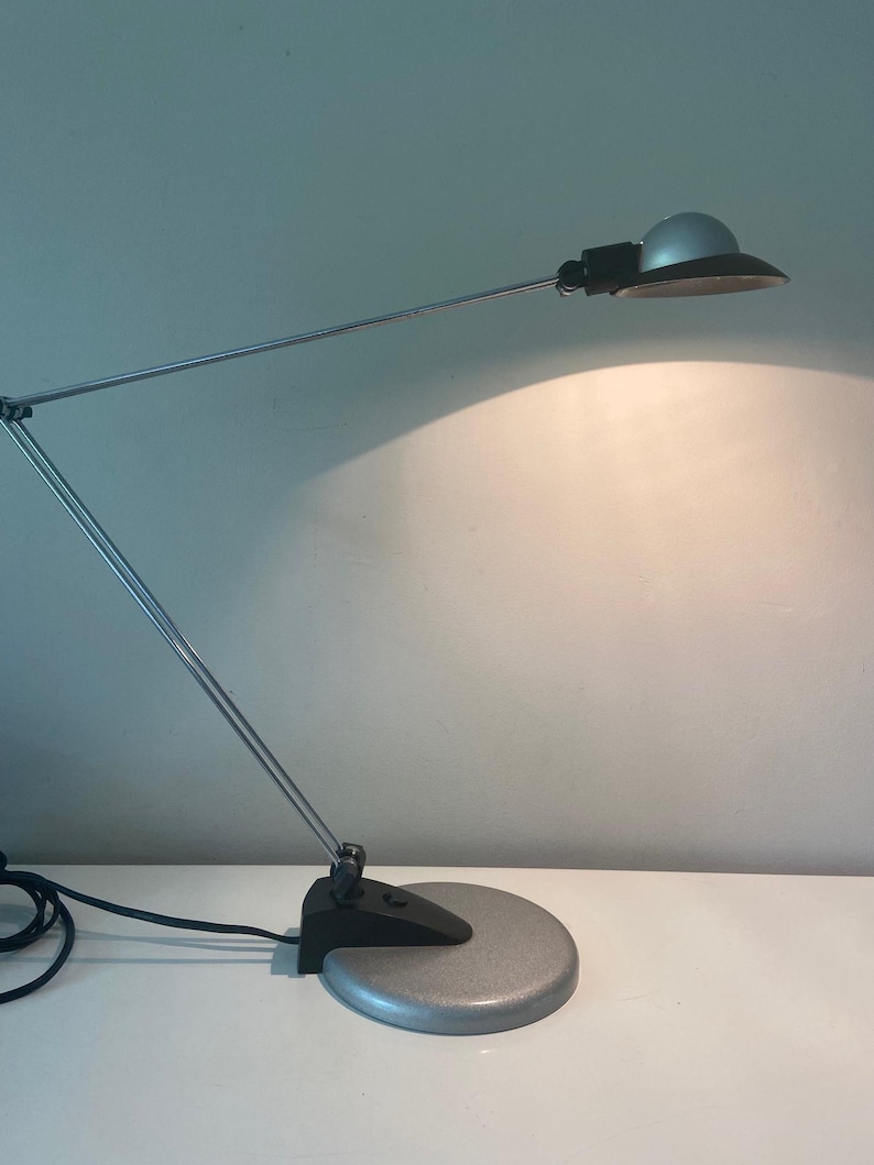 May include: A silver and black desk lamp with a flexible arm and a round base. The lamp is on and casting a warm light.