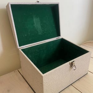 Pode incluir: Uma caixa de armazenamento retangular com um exterior texturizado em bege claro e um interior forrado com veludo verde vibrante. A caixa possui uma tampa articulada que está aberta e um mecanismo de fecho em metal prateado na parte frontal.