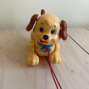 Op de afbeelding: Een gele plastic speelgoedhond met bruine oren en een rood trekkoord. De hond heeft een blauwe halsband en een lachend gezicht. Het speelgoed is ontworpen voor jonge kinderen.