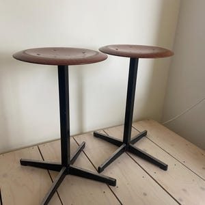 May include: Two tall stools featuring round, dark brown wooden seats with visible grain patterns. Each stool is supported by a central black metal post and a four-pronged black metal base, providing a stable foundation for the elevated seating surface.