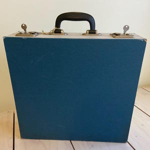 Pode incluir: Uma mala vintage azul-petróleo com uma alça preta e ferragens de metal. A mala tem duas chaves e um exterior texturizado. A mala está sobre uma superfície de madeira clara.
